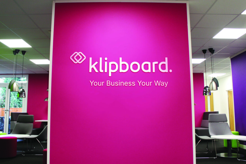 klipboard logo with "Your Business Your Way" text written underneath, all in white text on pink painted wall in office